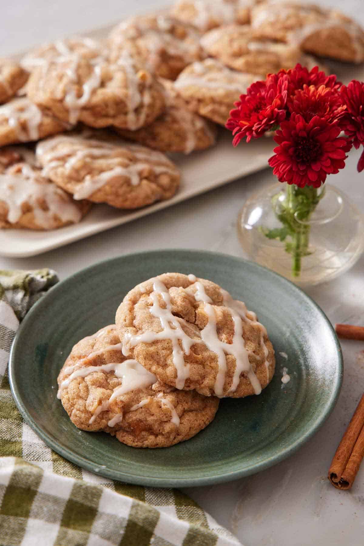 A plate with two brown butter wassail cookies with a platter more in the background along with some red flowers in a small vase.