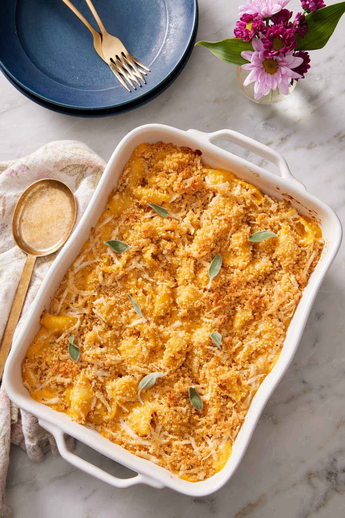 Overhead view of a baking dish of butternut squash mac and cheese. A serving spoon, flowers, plates, and forks on the side.