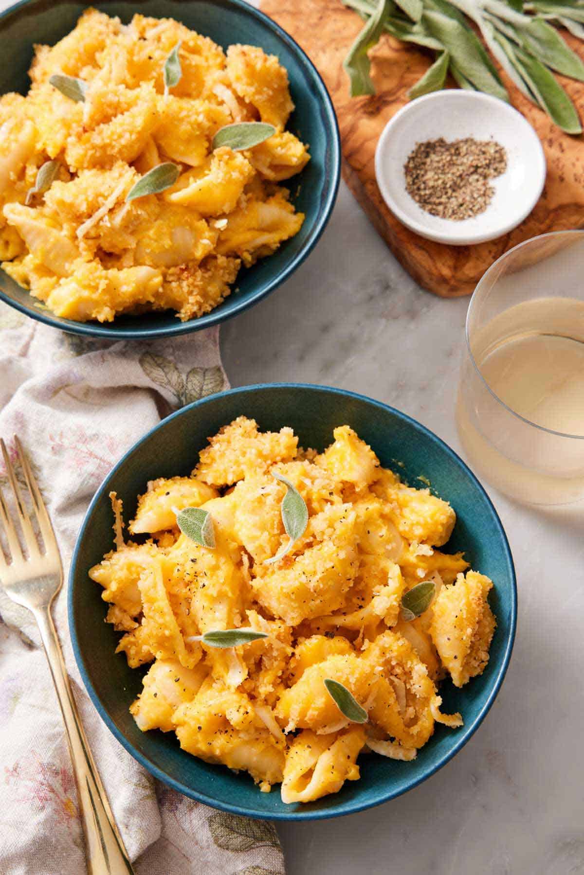 Two bowls of butternut squash mac and cheese with sage on top. A fork, a drink, a bowl of pepper, and more sage on the side.