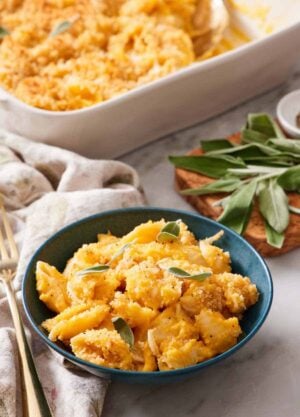 A bowl of butternut squash mac and cheese topped with sage. More fresh sage in the background along with the baking dish with the rest of the mac and cheese.