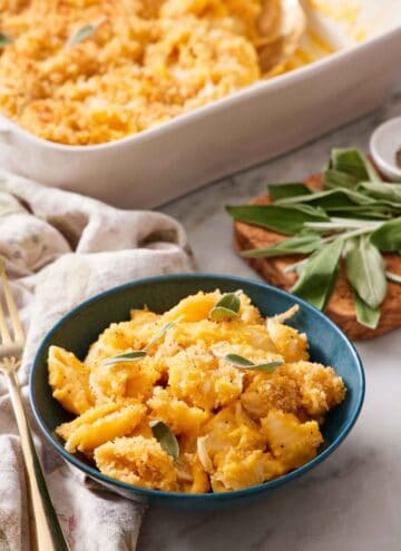 A bowl of butternut squash mac and cheese topped with sage. More fresh sage in the background along with the baking dish with the rest of the mac and cheese.