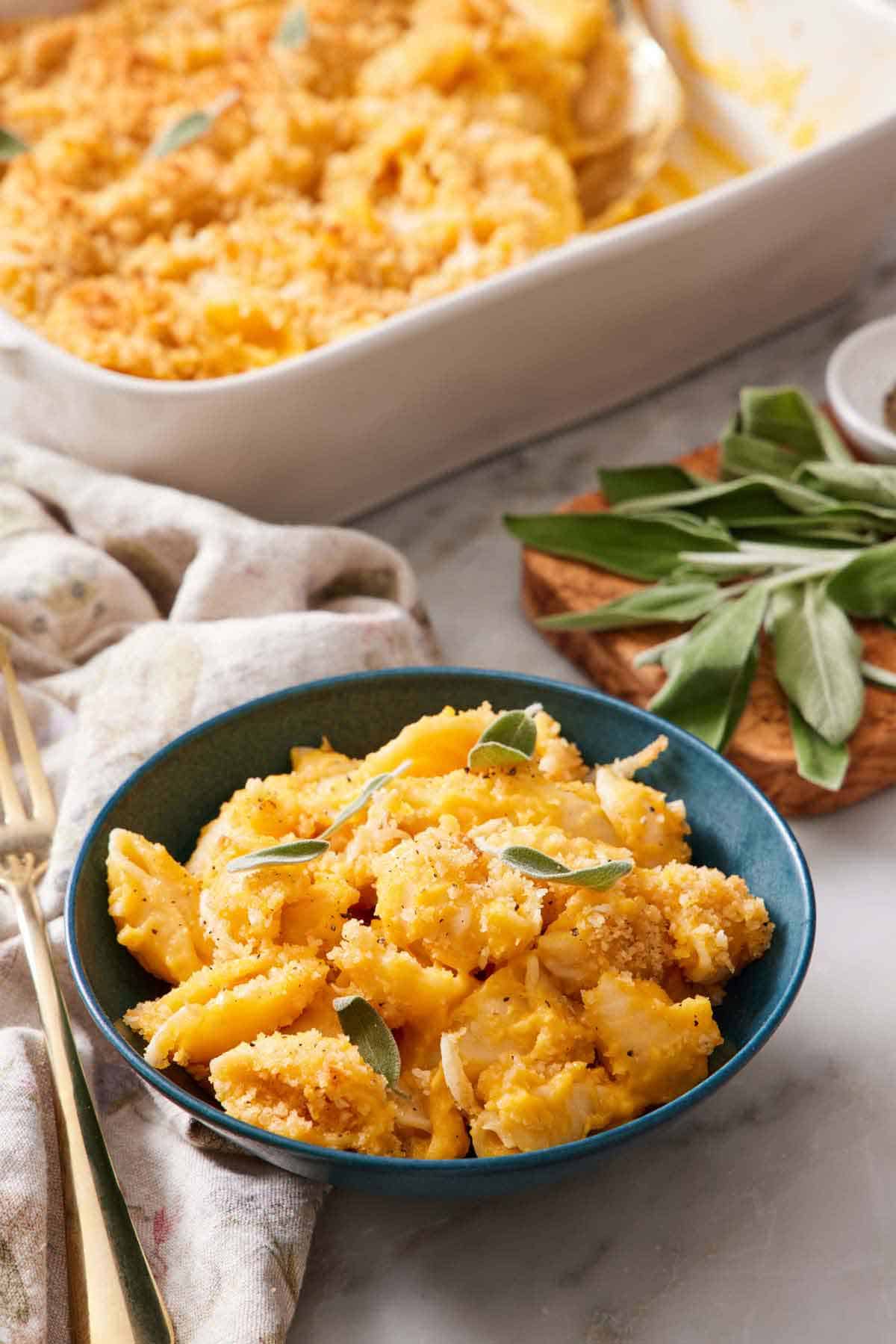 A bowl of butternut squash mac and cheese topped with sage. More fresh sage in the background along with the baking dish with the rest of the mac and cheese.