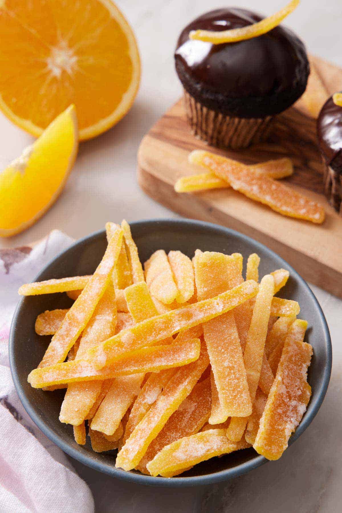 A bowl of candied orange peels. A chocolate glazed cupcake in the background with an candied orange peel on top along with cut oranges.