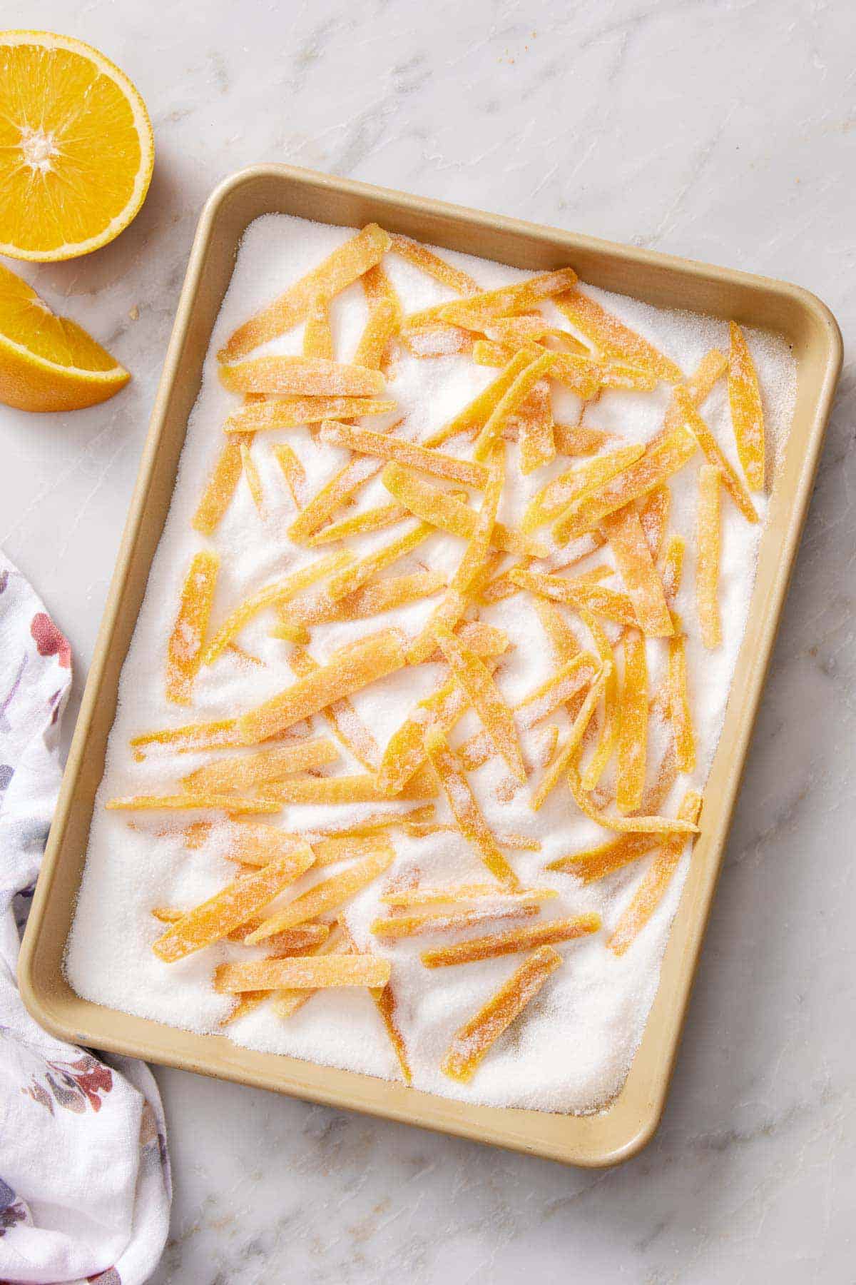 Overhead view of candied orange peels in a sheet pan of sugar. A cut orange off to the side.