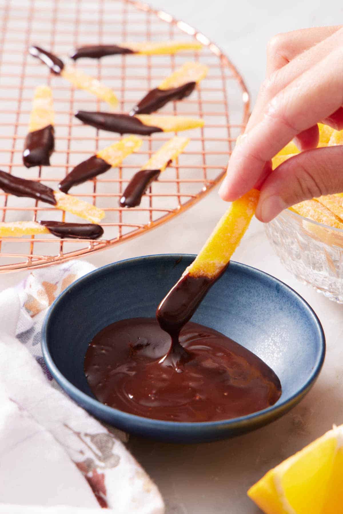 A candied orange peel dipped into a bowl of melted chocolate. More half dipped peels in the background on a cooling rack.