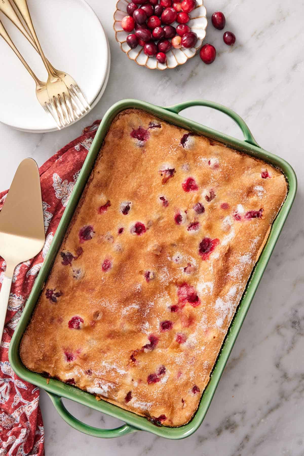 Overhead view of a green baking dish containing a cranberry cake. A bowl of fresh cranberries, a stack of plates, and forks on the side.