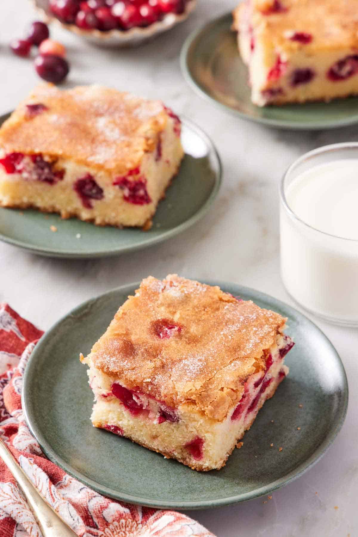 A plate with a square slice of cranberry cake with two more plated slices in the background along with a glass of milk.