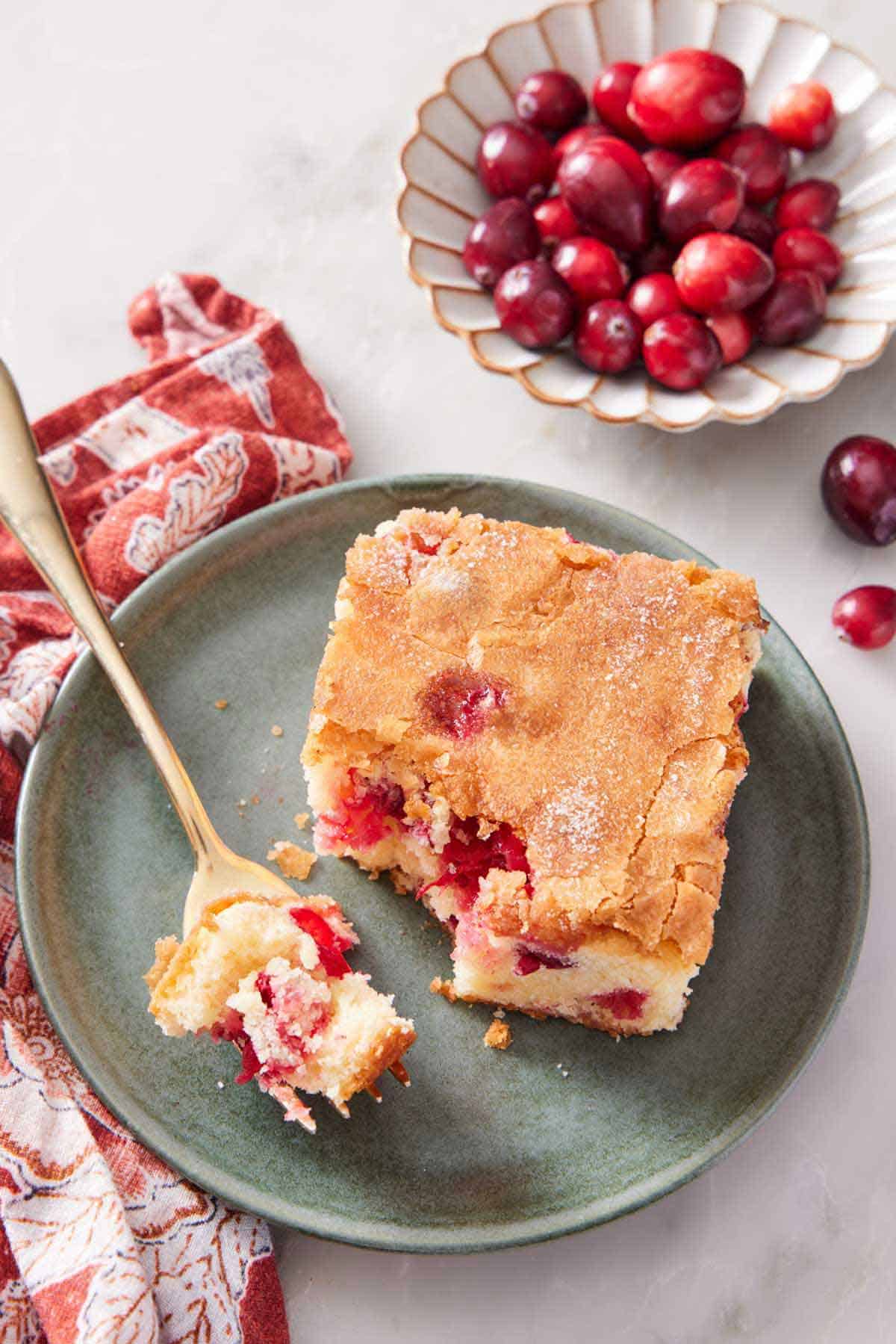 A plate with a slice of cranberry cake with a bite on a fork. A plate of fresh cranberries in the background.