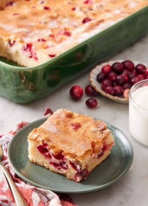 A plate with a square slice of cranberry cake with the rest of the cake in the background in a baking dish along with a glass of milk and fresh cranberries.