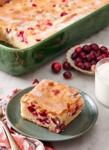 A plate with a square slice of cranberry cake with the rest of the cake in the background in a baking dish along with a glass of milk and fresh cranberries.