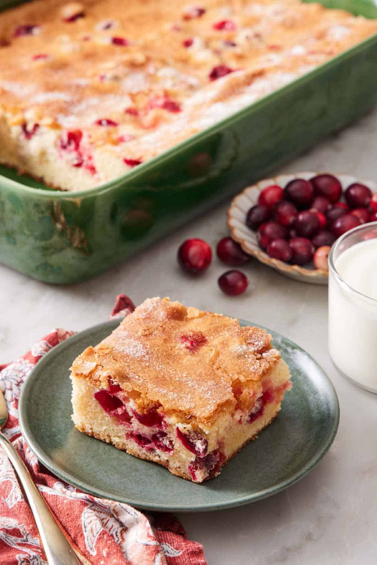 A plate with a square slice of cranberry cake with the rest of the cake in the background in a baking dish along with a glass of milk and fresh cranberries.