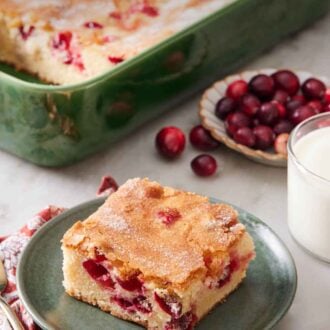 Pinterest graphic of a plate with a square slice of cranberry cake with the rest of the cake in the background in a baking dish along with a glass of milk and fresh cranberries.