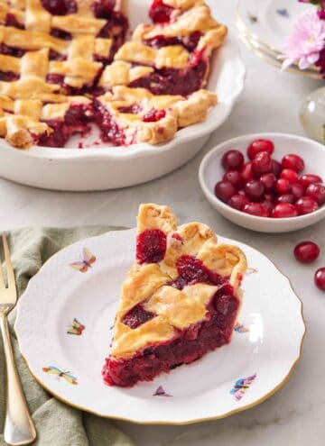 A slice of cranberry pie on a plate with a bowl of fresh cranberries and the rest of the pie in the background.