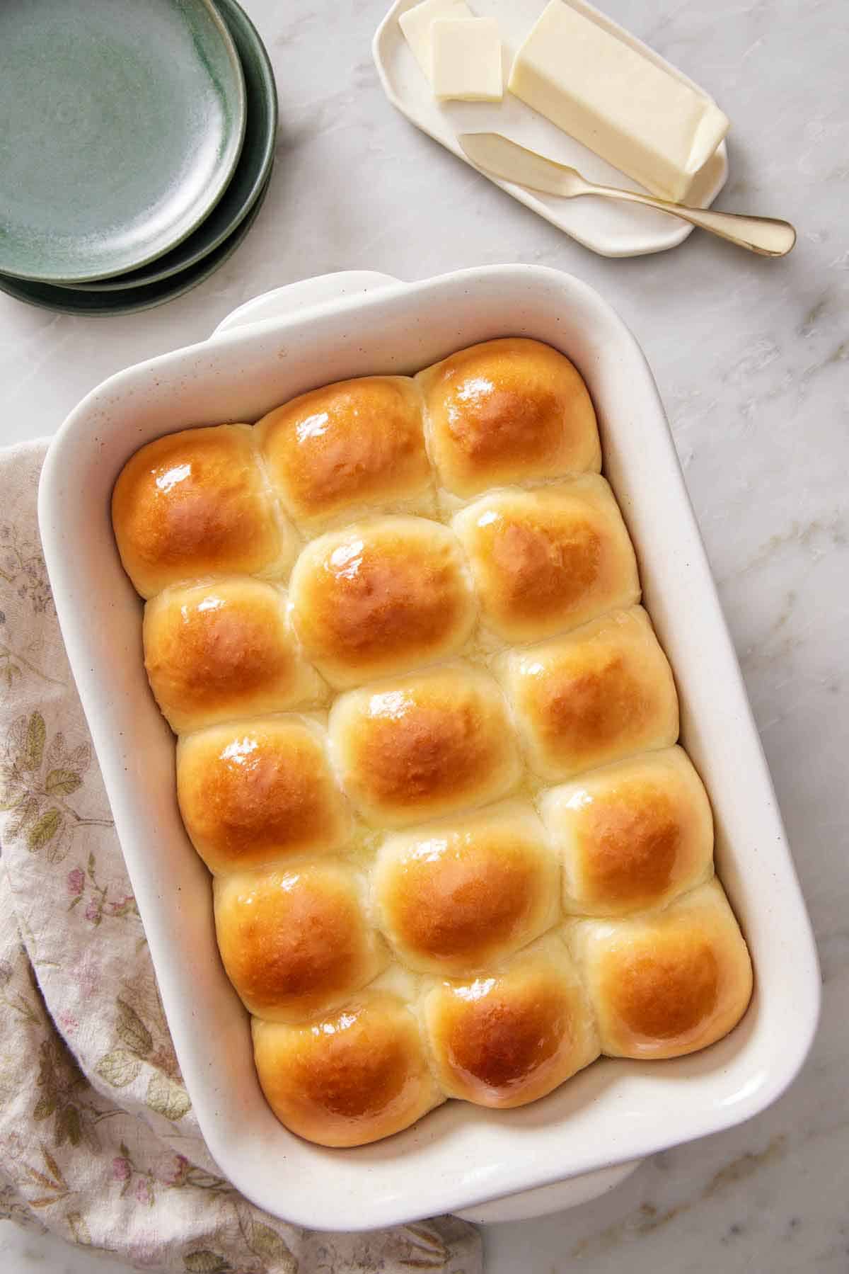 Overhead view of a white baking dish full of dinner rolls. A plate of butter and some green plates on the side.