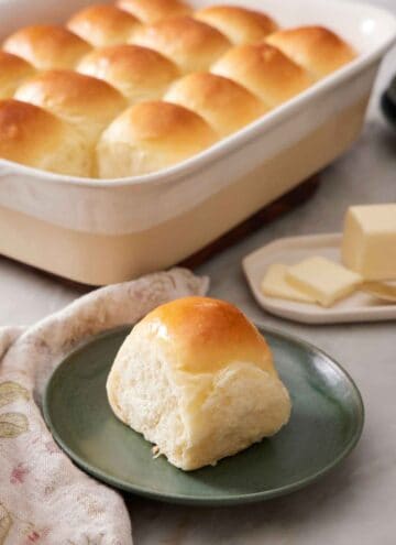 A green plate with a fluffy dinner roll with the rest of the baking dish in the background containing the rest of the rolls.