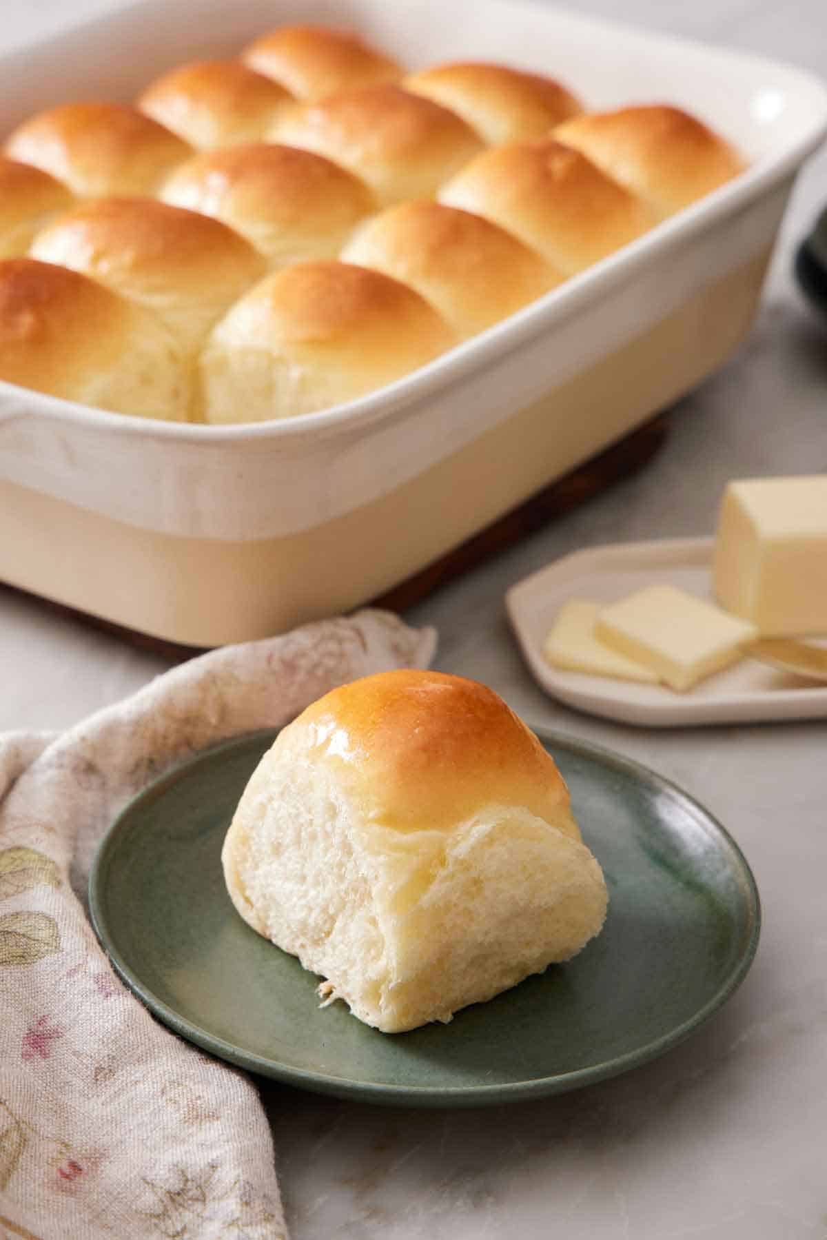 A green plate with a fluffy dinner roll with the rest of the baking dish in the background containing the rest of the rolls.