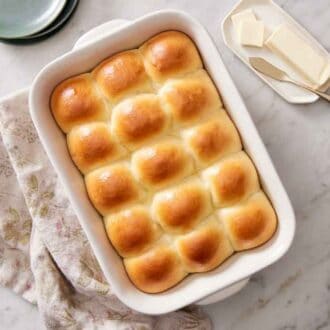 Overhead view of a white baking dish of golden dinner rolls. Butter, plates, and a napkin on the side.