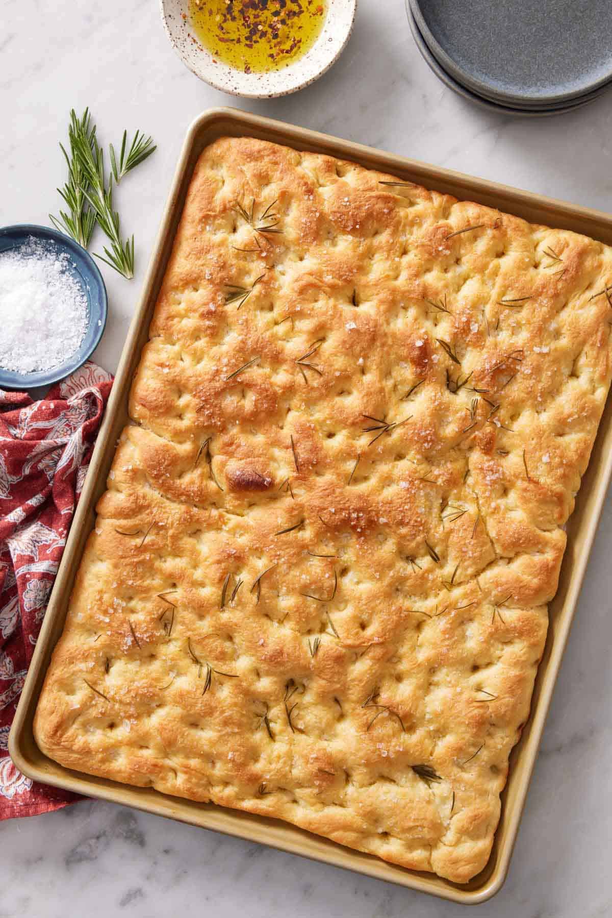 Overhead view of a sheet pan of focaccia topped with rosemary. A bowl of salt, dipping oil, and some rosemary on the side.