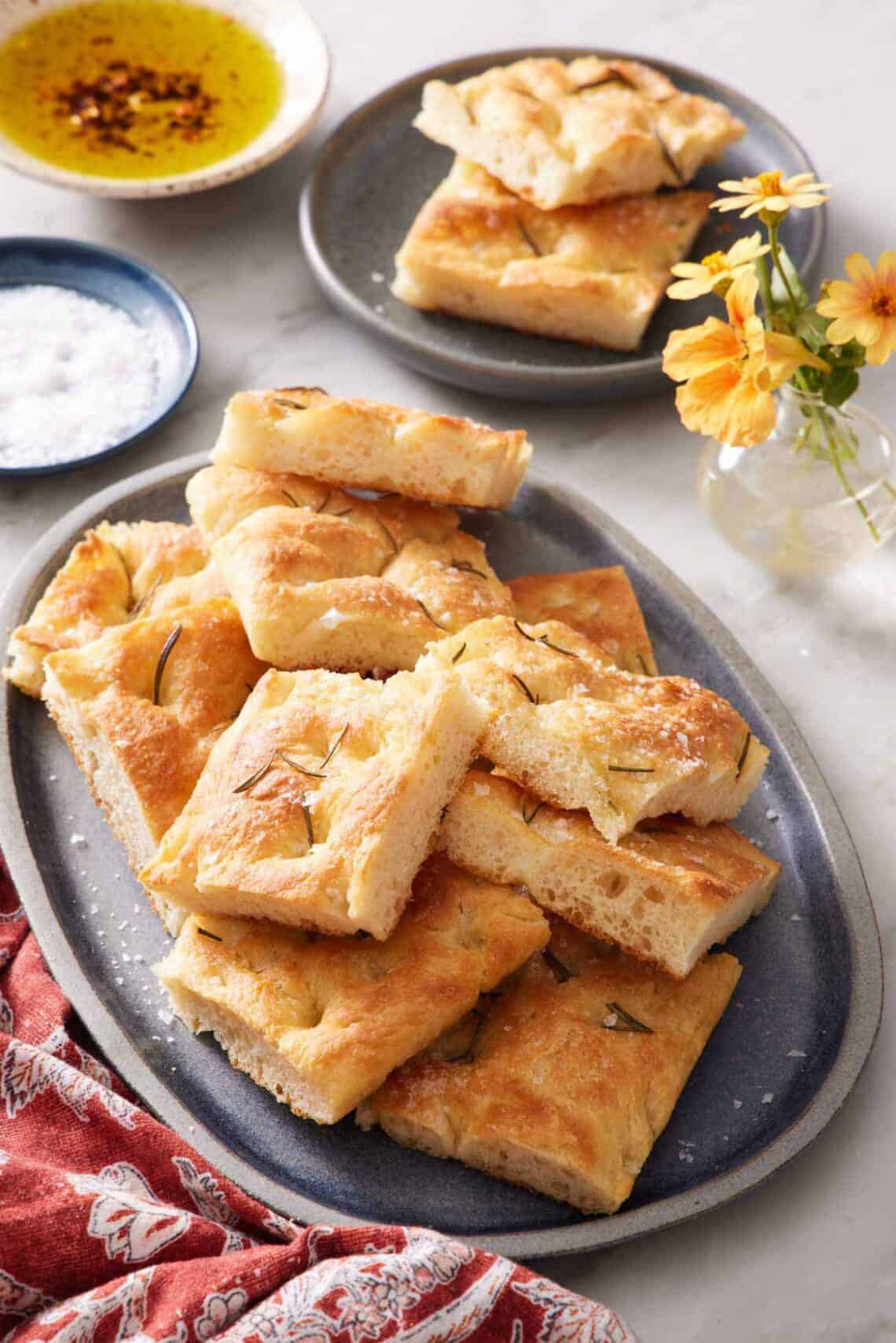 An oval platter with pieces of focaccia. A small vase of flowers, a plate with sliced focaccia, a bowl of oil, and a bowl of salt in the background.