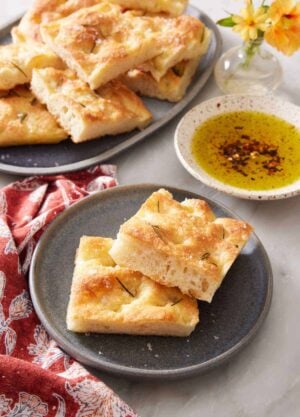 A plate with two pieces of focaccia with more on a platter in the background along with a bowl of oil for dipping.