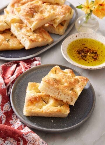 A plate with two pieces of focaccia with more on a platter in the background along with a bowl of oil for dipping.