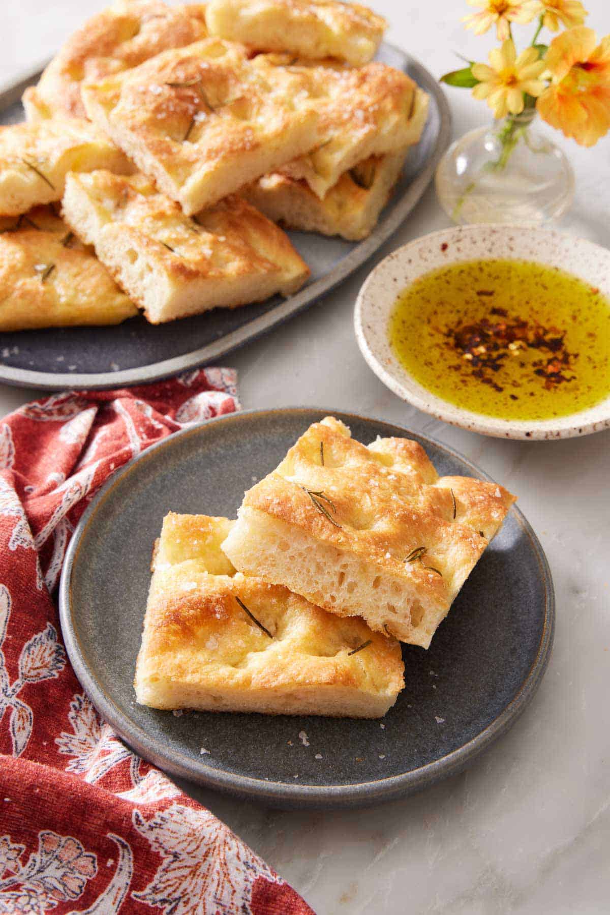 A plate with two pieces of focaccia with more on a platter in the background along with a bowl of oil for dipping.