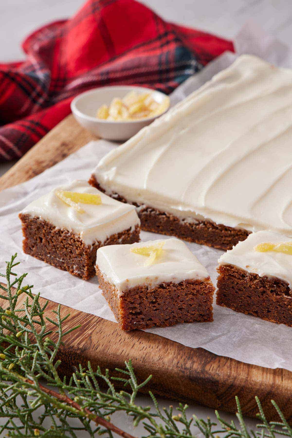 Gingerbread bars cut from the slab and topped with candied ginger on a parchment-lined cutting board with a bowl of candied ginger.