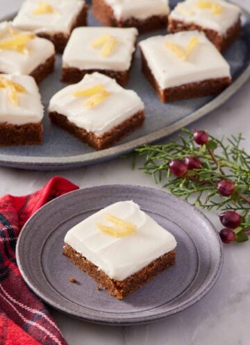 A plate with a gingerbread bar topped with candied ginger. A platter in the background with additional bars with some festive garnish on the side.