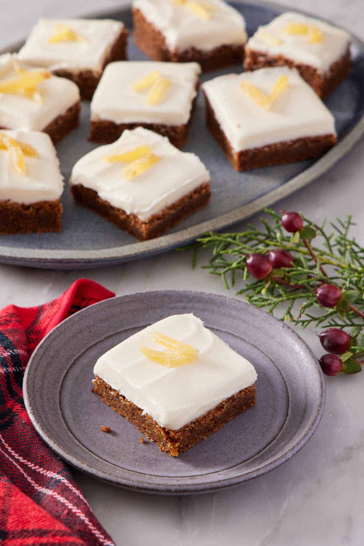 A plate with a gingerbread bar topped with candied ginger. A platter in the background with additional bars with some festive garnish on the side.