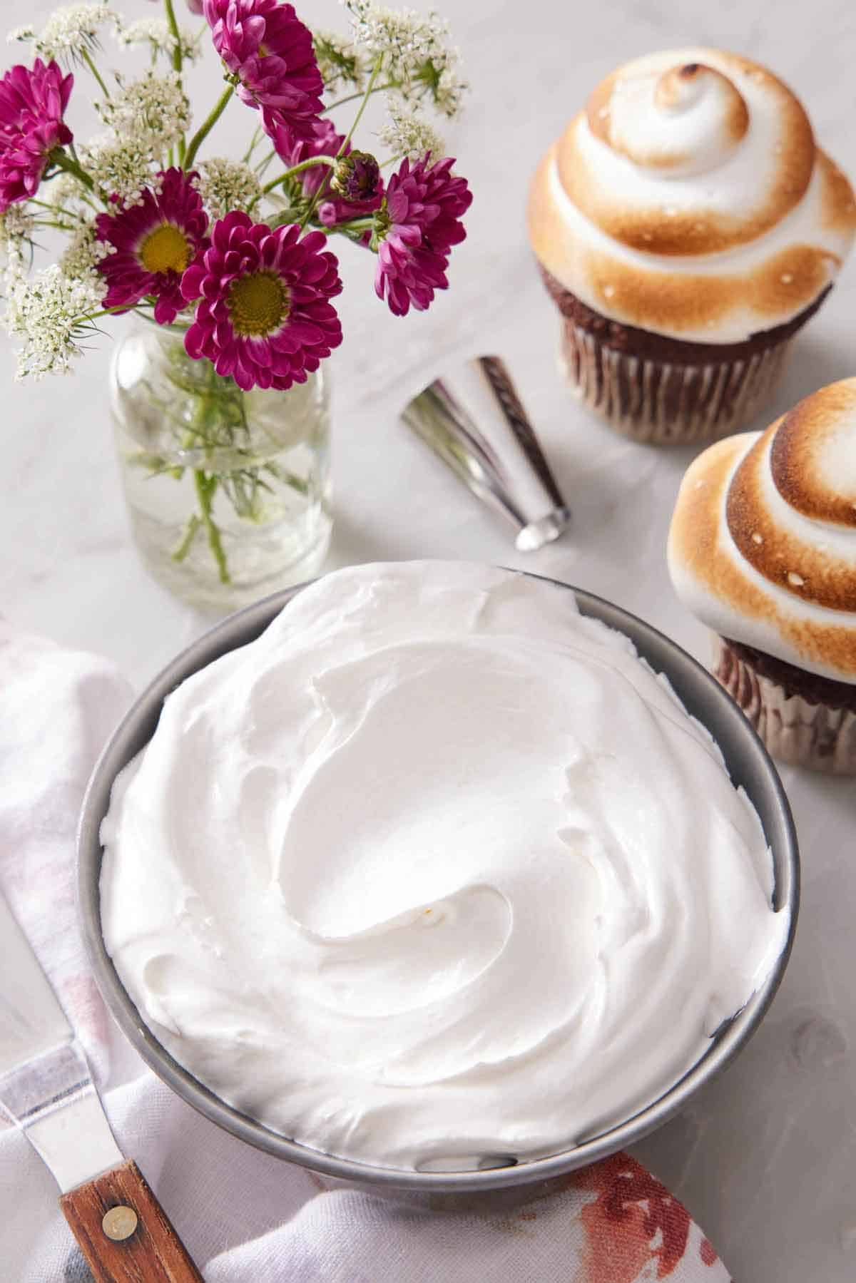 A bowl of homemade marshmallow frosting. A small vase of flower and two frosted and toasted cupcakes on the side.