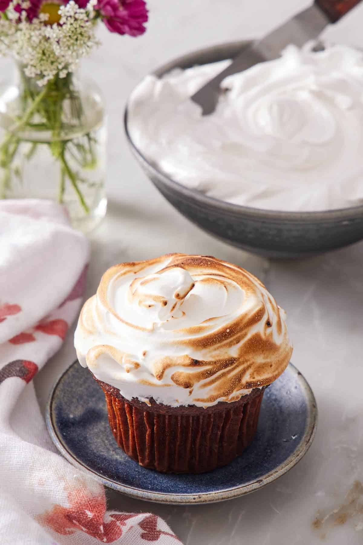 A cupcake topped with marshmallow frosting then toasted. A bowl of fluffy marshmallow frosting in the background along with some flowers.