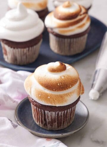 A chocolate cupcake topped with fluffy toasted marshmallow frosting. More frosted cupcakes in the background and a piping back on the side.