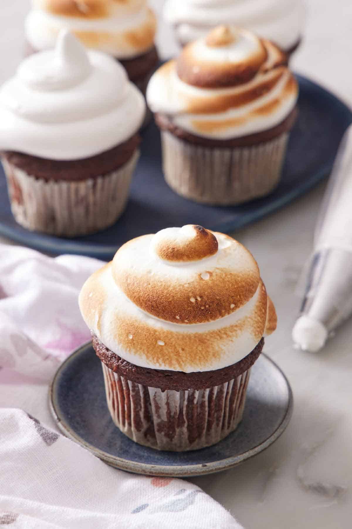 A chocolate cupcake topped with fluffy toasted marshmallow frosting. More frosted cupcakes in the background and a piping back on the side.