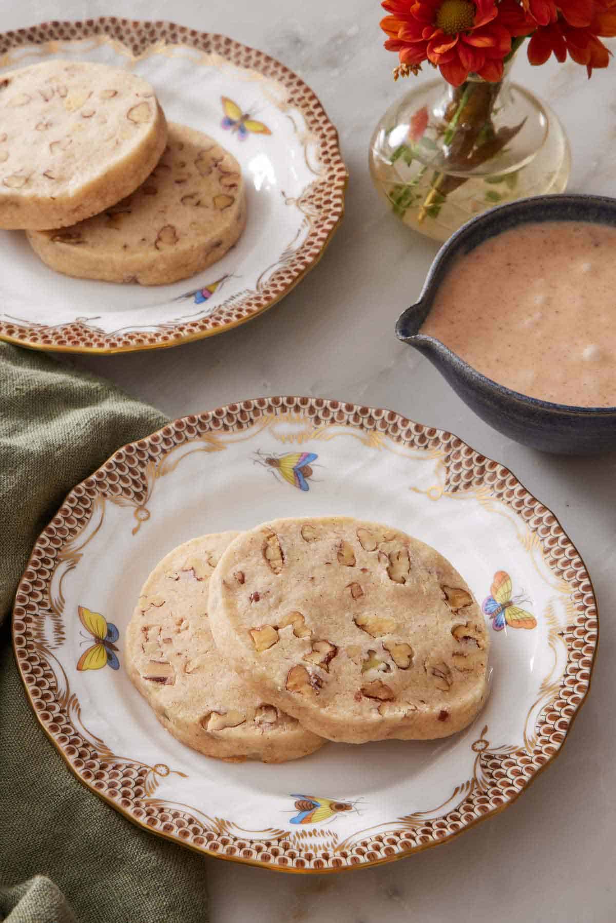 Two plates with two pecan shortbread cookies each. A bowl of glaze and a vase of flowers on the side.