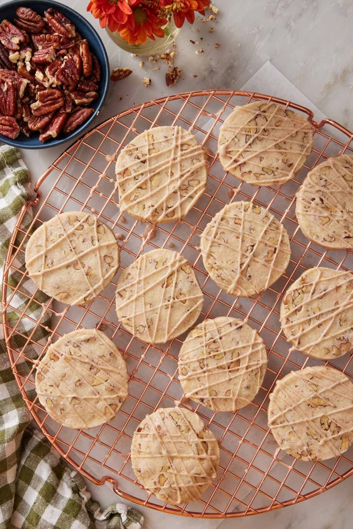 Overhead view of pecan shortbread cookies with glaze drizzled on top on a round cooling rack. A bowl of pecans on the side.