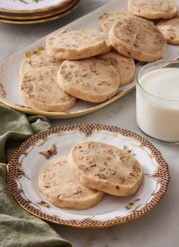 A plate with two pecan shortbread cookies with a glass of milk and platter of more cookies in the background.