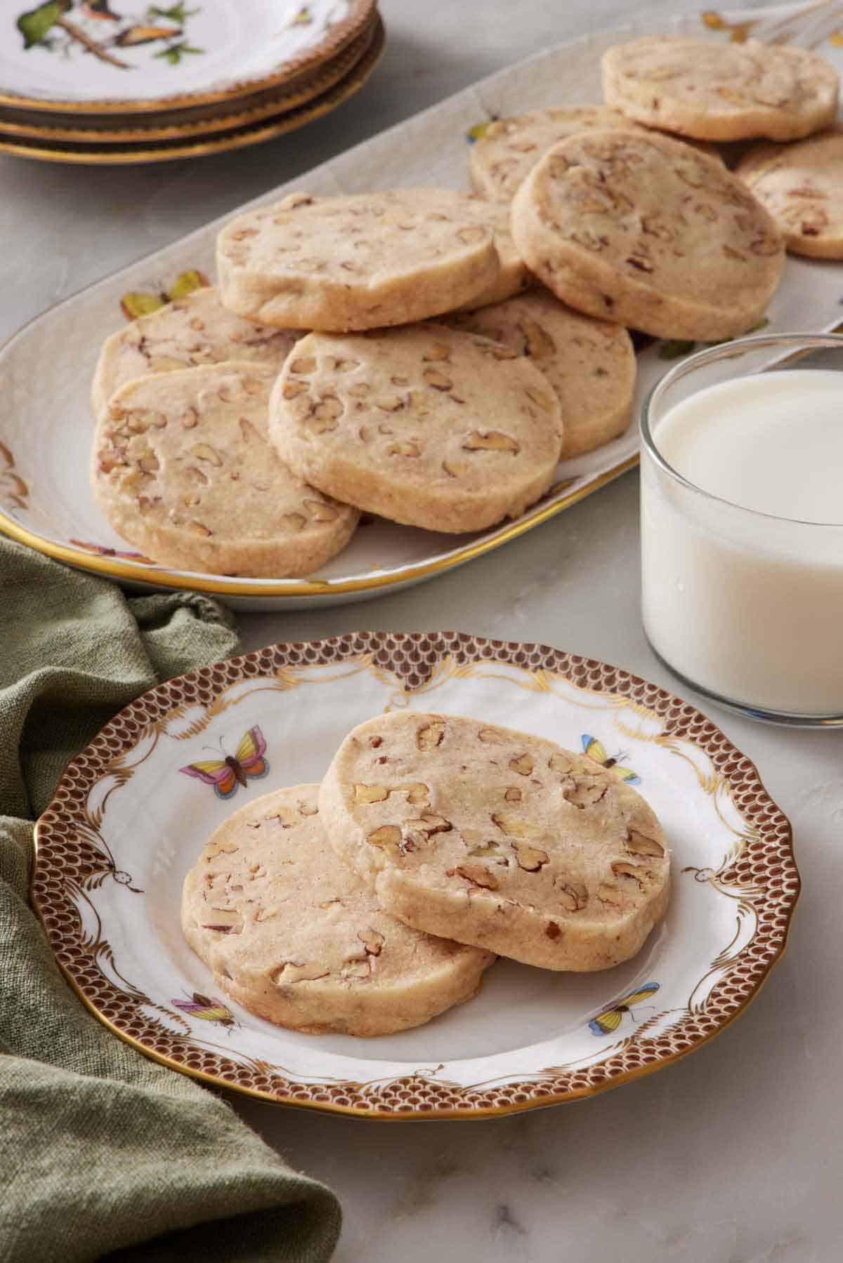 A plate with two pecan shortbread cookies with a glass of milk and platter of more cookies in the background.