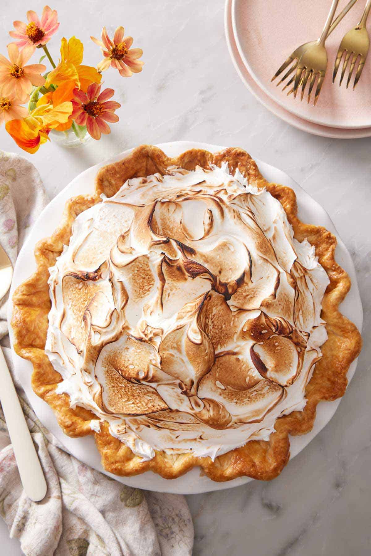 Overhead view of a pumpkin meringue pie with flowers, forks, and a stack of plates beside it.