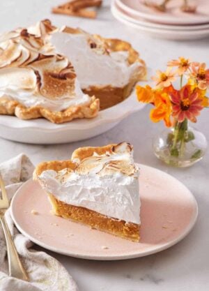 A plate with a slice of pumpkin meringue pie with flowers in the background along with the rest of the pie.