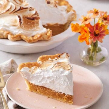 A plate with a slice of pumpkin meringue pie with flowers in the background along with the rest of the pie.