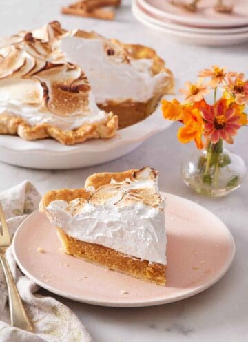 A plate with a slice of pumpkin meringue pie with flowers in the background along with the rest of the pie.