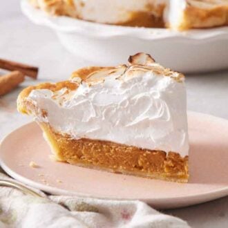 A slightly angled profile view of a slice of pumpkin meringue pie on a plate with a fork, flowers, cinnamon sticks, and the rest of the pie off to the side.