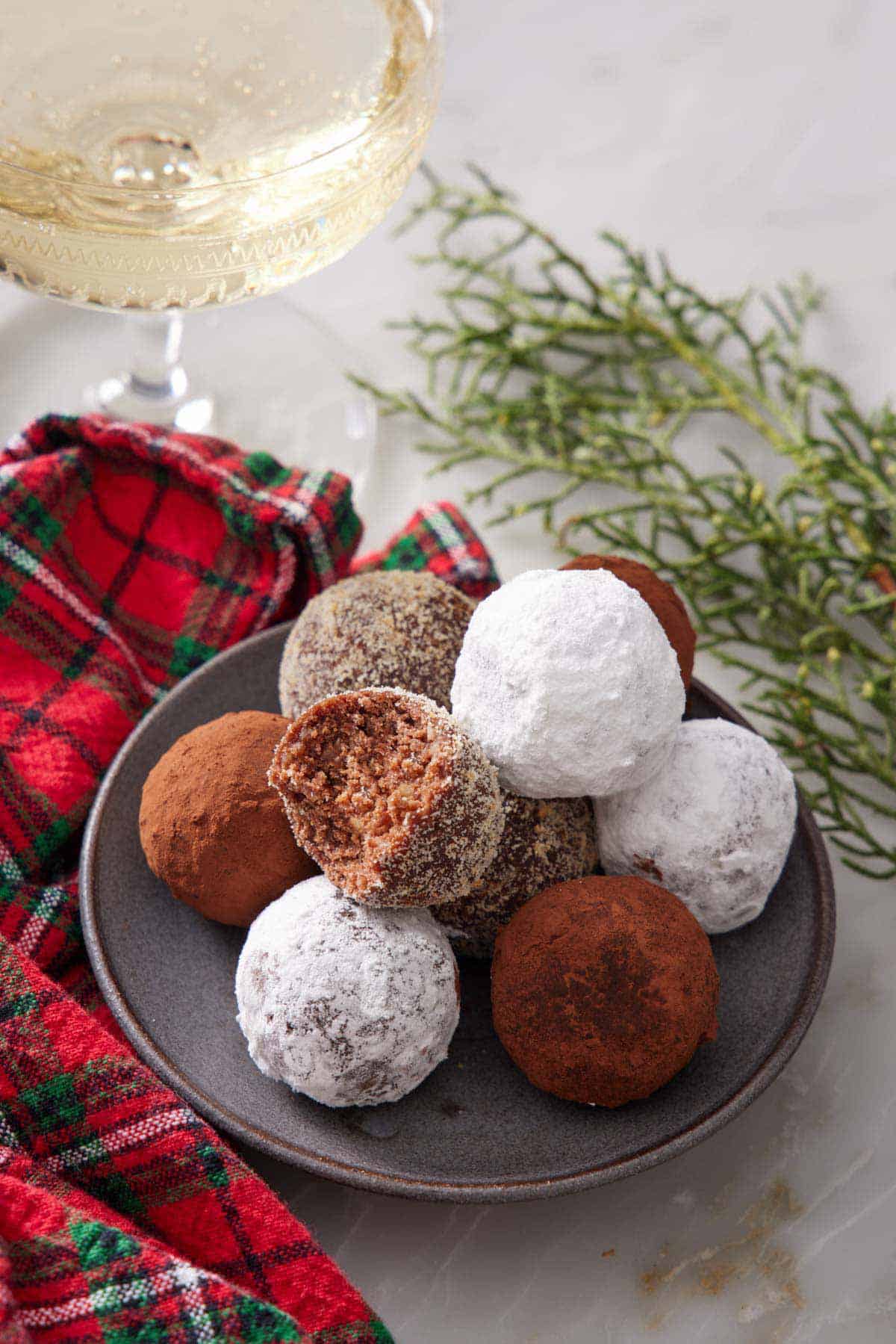 A plate with a pile of rum balls with one with a bite taken out of it. A glass of wine and some green garnish in the background.