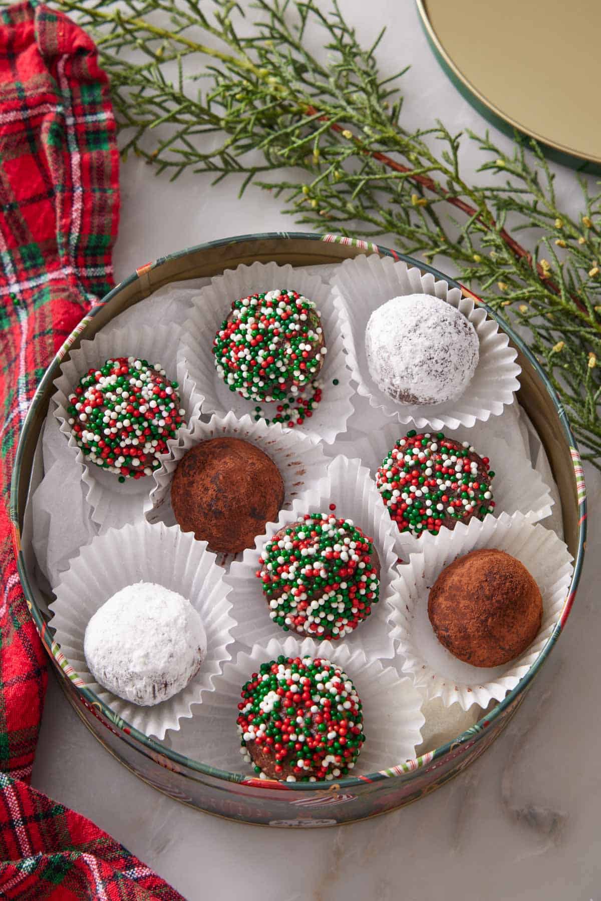 A round tin with rum balls in individual paper liners surrounded by a linen napkin and greenery. Rum balls coated in various toppings.