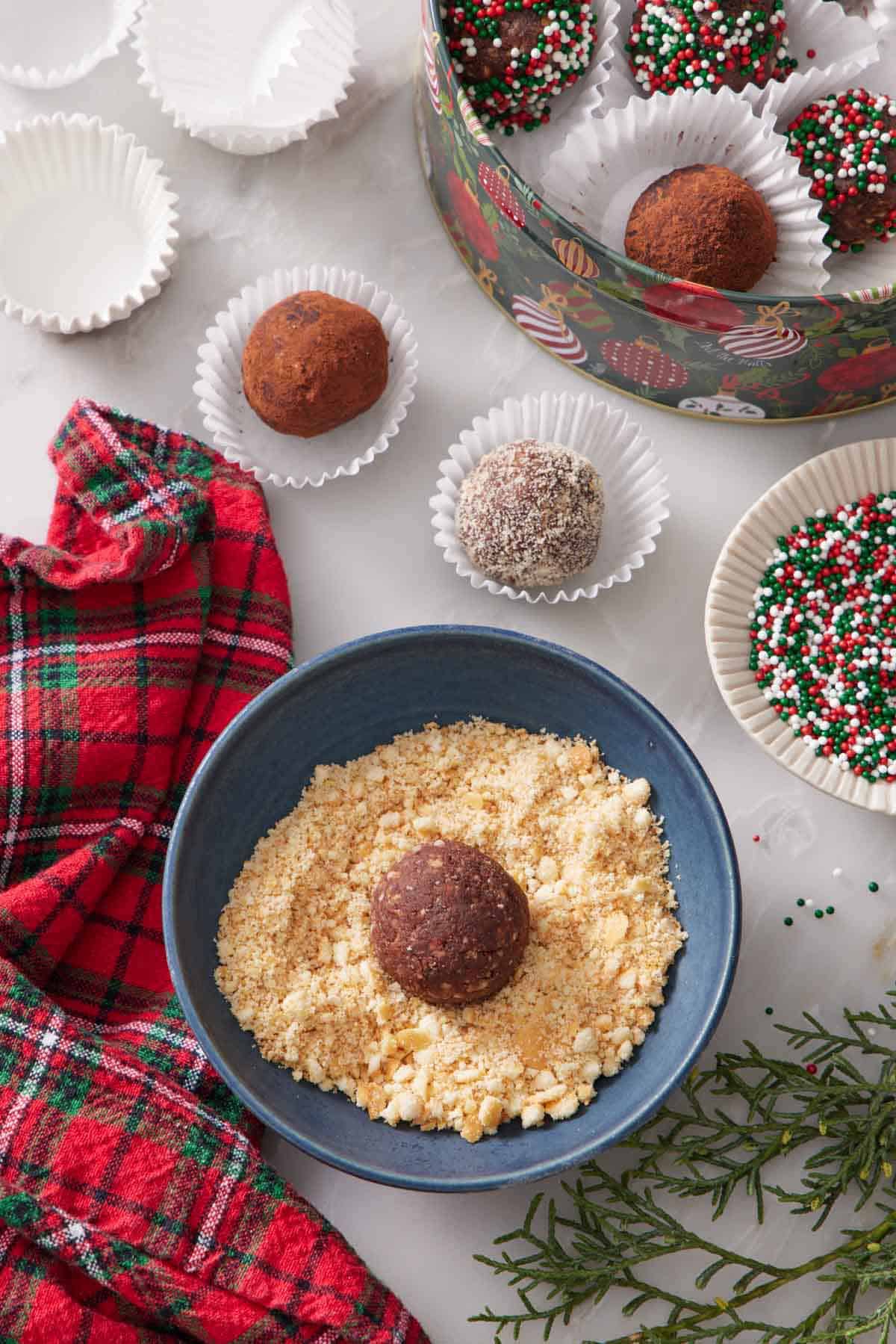 A bowl with finely chopped nuts with a rum ball in the middle. More rum balls in paper liners in the background with different coatings and a bowl of sprinkles.