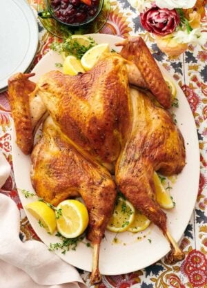 Overhead view of a platter with a spatchcock turkey surrounded by fresh herbs and lemons. A bowl of cranberry sauce and plates off to the side.