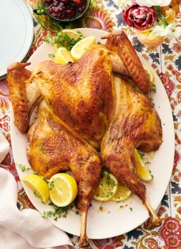 Overhead view of a platter with a spatchcock turkey surrounded by fresh herbs and lemons. A bowl of cranberry sauce and plates off to the side.