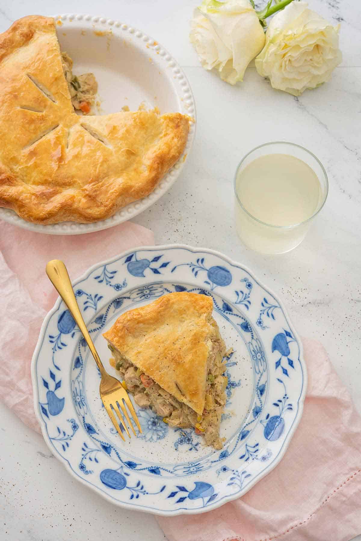 Overhead view of a slice of turkey pot pie on a plate with a fork. A drink and the rest of the pie off to the side.