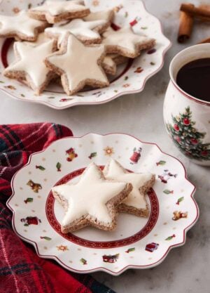 A plate with two star shaped zimtsterne with more in the background on a larger platter. A coffee beside it.