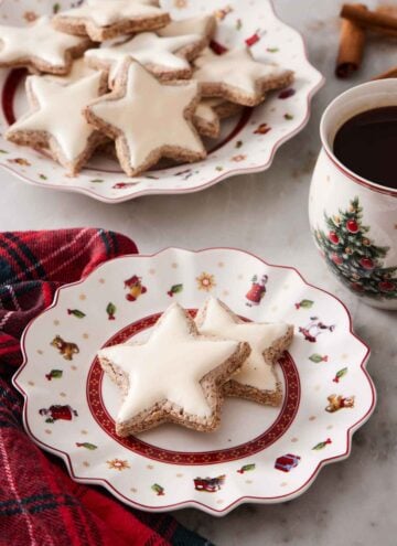 A plate with two star shaped zimtsterne with more in the background on a larger platter. A coffee beside it.